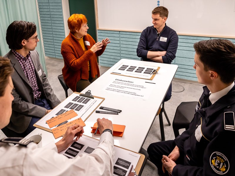 The photos shows five persons, sitting in a round and discussing.