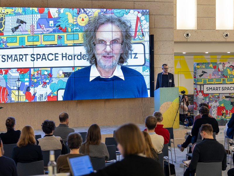 Das Foto zeigt Oliver Schruoffeneger in einem Video auf einer Leinwand, davor sitzen die Teilnehmer:innen der Veranstaltung.