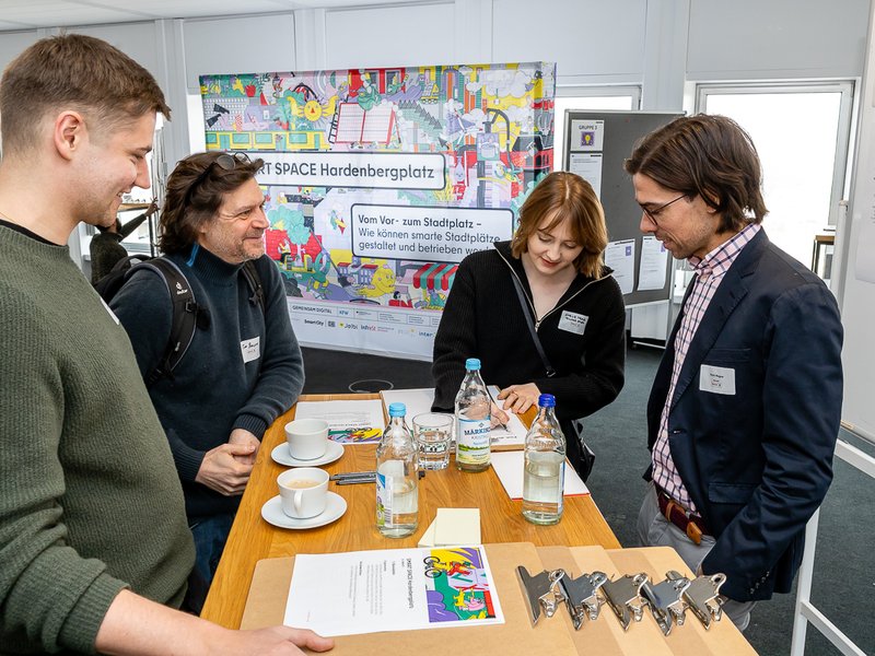 The photos shows four persons, standing together around a table as a group and discuss.