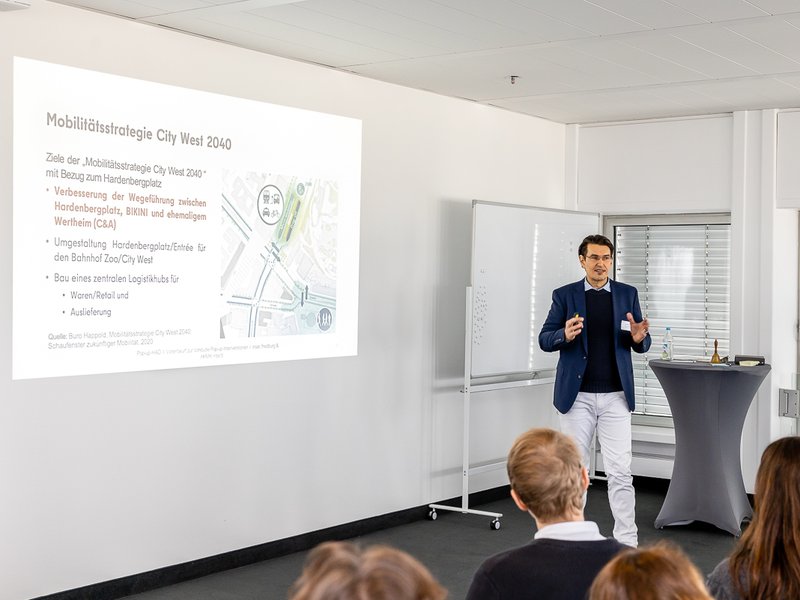 The photo shows Rolf Mienkus, holding a speech, In the background you can see a presentation slide about the mobility strategie for City West 2040.