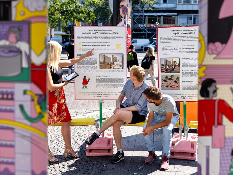 Frau zeigt auf Tafeln der Ausstellung, während zwei Personen auf den Bänken sitzen.