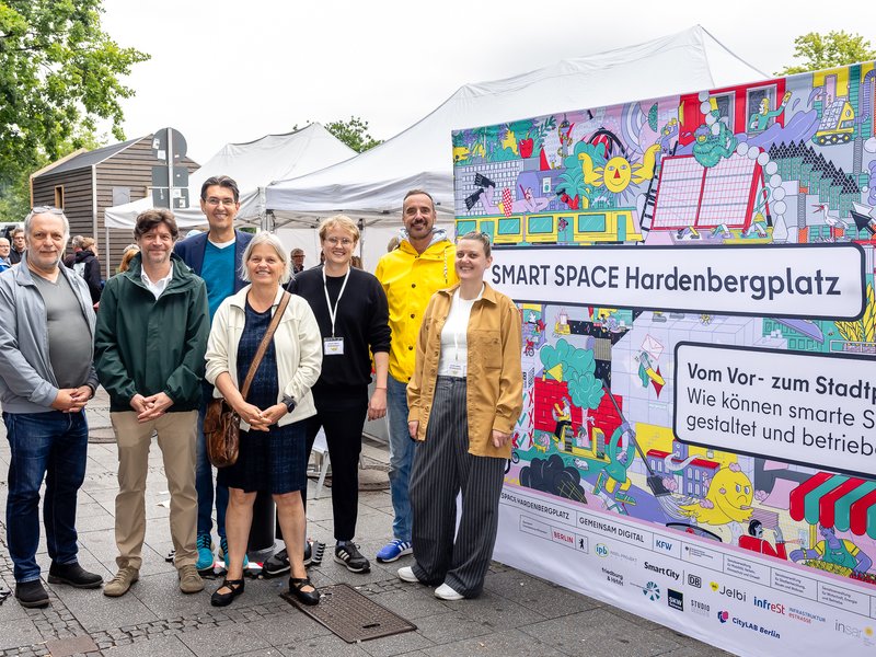 Ein Bild von dem Team vor der bunten Messewand des Pilotprojekts SMART SPACE Hardenbergplatz.